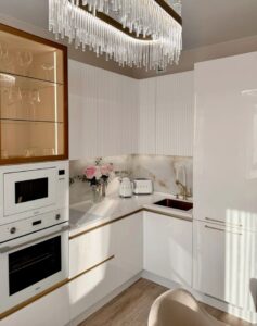 آشپزخانهای مدرن با کابینتهای سفید براق، دستگیرههای طلایی، بین کابینتی سنگی با رگههای طلایی، نورپردازی کریستالی و ترکیب چوب در طراحی، فضایی شیک و دلنشین را به نمایش میگذارد.